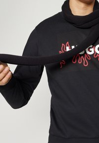 Maglione nero con colletto alto e design di fiamme rosse attorno al logo. Il modello tiene una sciarpa spessa coordinata.