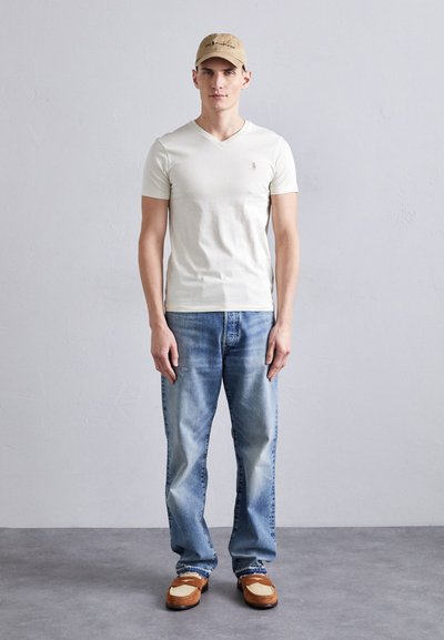 Chemise blanche à manches courtes en col en V, jeans délavés bleu clair, mocassins marron et beige, casquette beige avec texte. Se tenant contre un fond neutre.