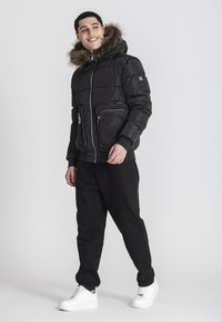 Giacca a vento nera con cappuccio foderato in pelliccia, tasche con zip e polsini a costine, abbinata a pantaloni neri e sneakers bianche.