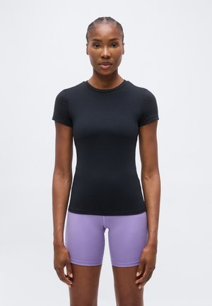 Vrouw met gevlochten haar, gekleed in een aansluitend zwart T-shirt met korte mouwen en lavendelkleurige fietsshorts, staand tegen een effen grijze achtergrond.