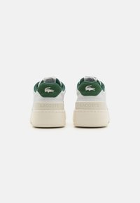 White sneakers with green accents, textured back, padded collar, and raised sole featuring the Lacoste logo prominently displayed.