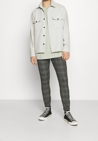 Ljust grå knapp-up jacka med två bröstfickor, matchad med en grön t-shirt och grå rutig slim-fit byxor, kompletterad med svarta hög-top sneakers.