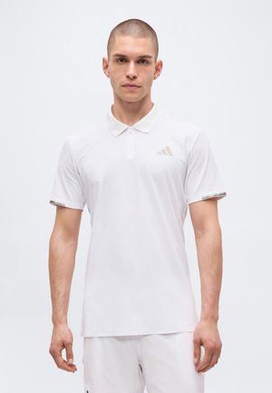 Bărbat cu păr scurt, purtând un tricou polo alb cu mâneci scurte și pantaloni albi, stând în picioare în fața unui fundal simplu și luminos.
