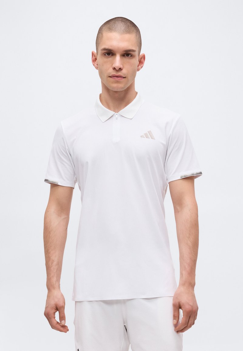 Homme aux cheveux courts portant un polo blanc à manches courtes et un pantalon blanc, debout devant un fond clair uni.