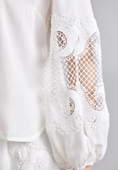 Vit blus med intrikata spetsärmar med broderade mönster, ett nätmönster och delikata pärlaccents. Mjuk, lättviktsstoff.