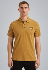 Hořčicově žlutá polo košile s tříknoflíkovou légou, krátkými rukávy a potiskem značky na hrudi. Vyrobena z měkké látky.