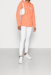 Felpa corallo con scritta bianca, abbinata a jeans skinny bianchi e sneakers bianche. L'outfit include una piccola borsa a tracolla bianca.