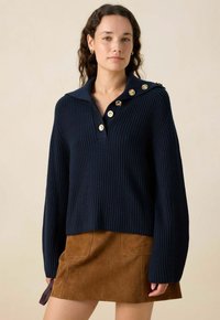 Pull en maille côtelée navy avec un col, agrémenté de boutons dorés sur l'épaule, associé à une mini-jupe en daim marron.