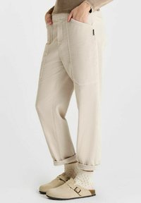 Beige corduroy broek met een relaxte pasvorm, voorzien van zijzakken en omgeslagen hemden, gecombineerd met lichtgekleurde instappers en geblokte sokken.