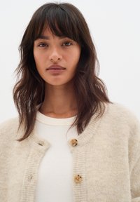 Cardigan en maille beige clair avec des bords côtelés et des boutons ronds ; porté sur un haut crème. Texture douce et coupe ample.