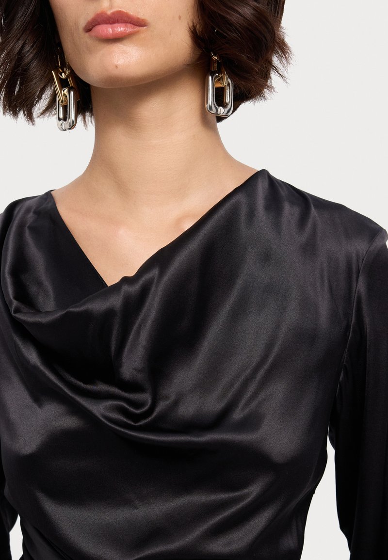 Top de satén negro con escote drapeado y mangas largas, que presenta pendientes brillantes en forma de cadenas plateadas y doradas. Textura suave, detalle mínimo.