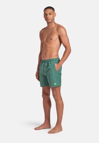 Uomo atletico in piedi a piedi nudi, senza maglia, con indosso pantaloni da bagno verdi con tasche, su uno sfondo bianco semplice.