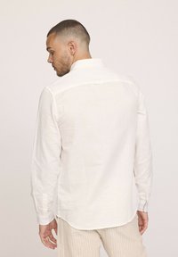 Homme aux cheveux courts portant une chemise blanche à manches longues et un pantalon beige à rayures, de dos contre un fond clair.