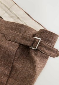 Cinturón de tela texturizada marrón con hebilla de metal plateado, sujeto a un par de pantalones con forro interior beige.
