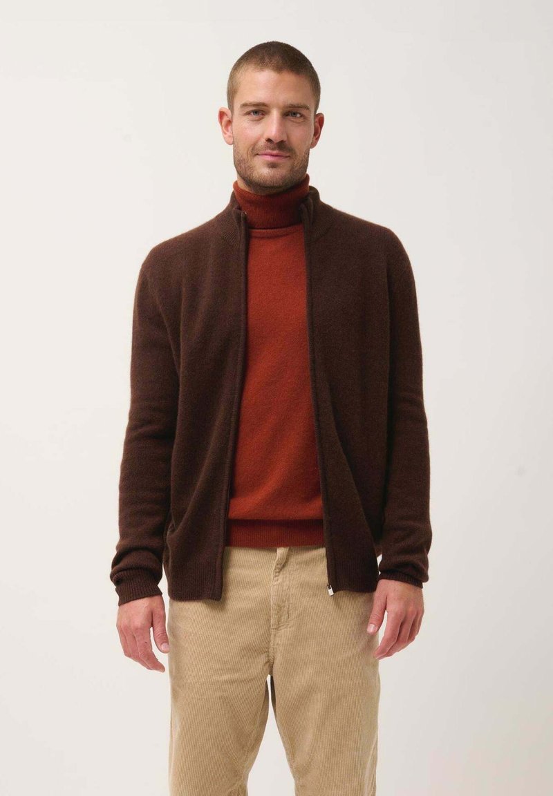 Cardigan marrone con zip sopra un dolcevita rosso, abbinato a pantaloni in corduroy beige. Tessuto liscio, polsini e colletto a coste.