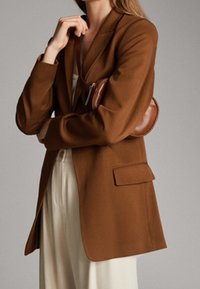 Brązowa marynarka o dopasowanym kroju, zapinana na jeden guzik, z klapową kieszenią. Modelka trzyma małą, okrągłą brązową torbę, nosi kremowe spodnie.