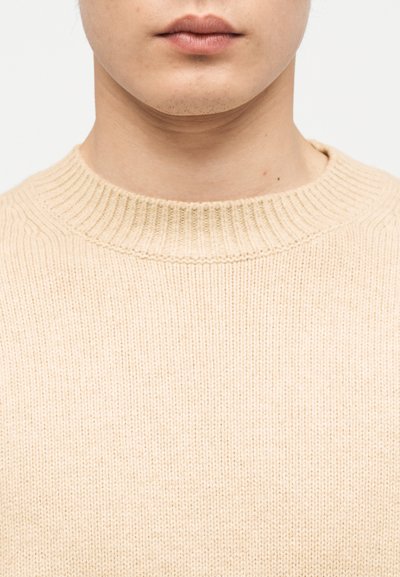 Gros plan sur le bas du visage et le cou d'une personne portant un pull en tricot côtelé beige sur un fond uni.