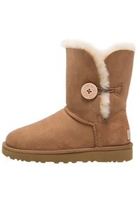 Bota de invierno de ante marrón con un suave forro de piel de oveja blanca, un botón redondo con la marca UGG en el lateral y una suela de goma plana.