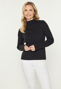 Schwarzer gerippter Rollkragenpullover mit langen Ärmeln, eng anliegendem Design und strukturierter Stoff. Kombiniert mit weißen Hosen.