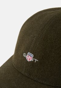 Oliwkowa wełniana czapka z zaokrągloną koroną, z haftowanym logo w białym i niebieskim kolorze. Miękka tekstura z widocznymi szwami.