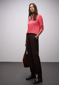 Top rosa de manga larga, pantalones anchos de pierna en marrón oscuro y botines de cuero negro. La modelo sostiene un bolso de ante marrón. Fondo sencillo.