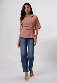 Top avvolgente rosa polveroso con maniche corte, dettaglio con fiocco in vita e cuciture bianche. Abbinato a jeans a gamba larga blu chiaro e sandali bianchi.