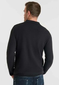 Uomo di spalle che indossa un maglione nero a maniche lunghe semplice e jeans blu, su uno sfondo chiaro.