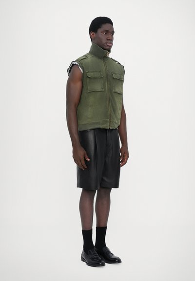 Gilet sans manches vert avec col montant, zip avant et deux poches poitrine. Porté avec des shorts en cuir noir, des chaussettes noires et des mocassins.