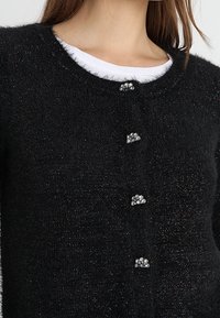 Musta pörröinen neuletakki pyöreällä pääntiellä, jossa on neljä koristeellista kristallipainiketta ja hienovarainen kimallus koko kankaassa.