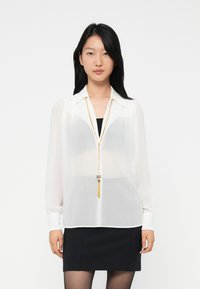 Young woman with long black hair wearing a sheer white blouse over a black top, black skirt, and a long gold necklace, standing against white background.