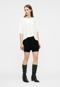 Γυναίκα που στέκεται φορώντας άνετο λευκό τοπ, μαύρα σορτς με πιέτες και μαύρες μπότες με τακούνι μέχρι το γόνατο, μπροστά σε απλό φόντο.