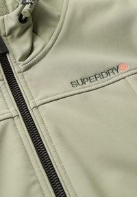 Lichtgroene ritsjack van gladde stof. Voorzien van een zwarte rits, gestikte accenten en een geborduurd "SUPERDRY" logo in donkergroen en oranje.