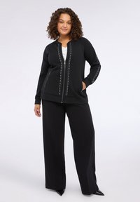 Felpa nera con zip e dettagli di borchie lungo la zip e le maniche, abbinata a pantaloni neri a gamba larga e scarpe nere a punta.