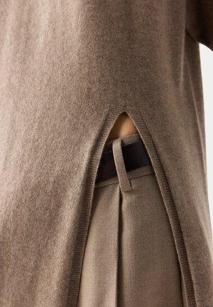 Brauner Strickpullover mit seitlichem Schlitz, der beigetöne Hose und schwarzen Gürtelriemen zeigt, wobei die Stoffstruktur und Kleidungsdetails sichtbar sind.