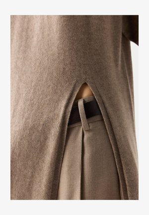 Brauner Strickpullover mit seitlichem Schlitz, der beigetöne Hose und schwarzen Gürtelriemen zeigt, wobei die Stoffstruktur und Kleidungsdetails sichtbar sind.