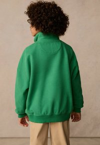 Zeleni oversized pulover s visokim ovratnikom in rebrastimi manšetami, izdelan iz mehkega materiala, s krožno jeko na hrbtu.