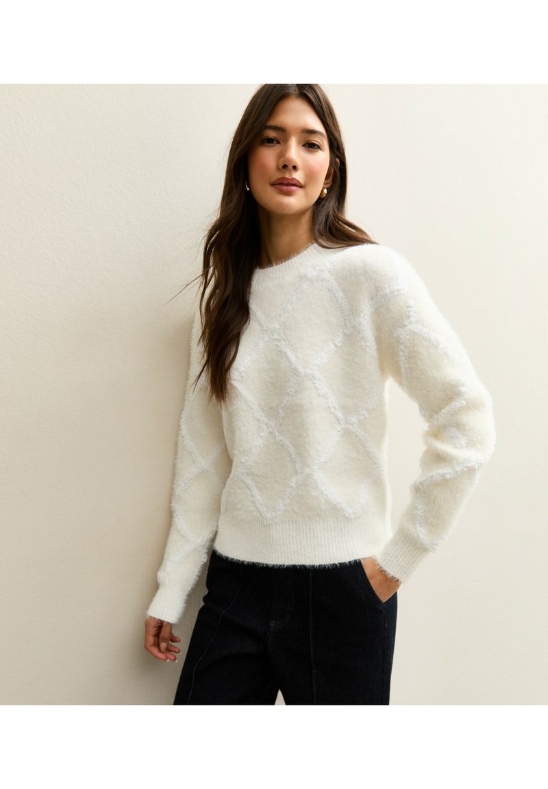 Maglione bianco e soffice con motivo a diamante, maniche lunghe, polsini e orlo a coste. Abbinato a pantaloni di jeans scuri a vita alta.