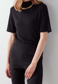 Žena oblečená v černém tričku s krátkým rukávem a odpovídajících kalhotách stojí proti světlé zdi a vystavuje jemné prsteny na svých prstech.