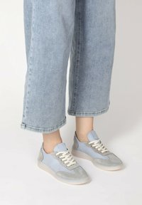 Jasnoniebieskie sneakersy z szarymi zamszowymi akcentami, okrągłym czubkiem, kremowymi sznurówkami i teksturowanym materiałem. Noszone z podwiniętymi, jasnoniebieskimi dżinsami.