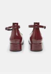 Red patent leather ankle strap shoes with a low block heel, featuring a smooth, shiny finish and minimalistic design.