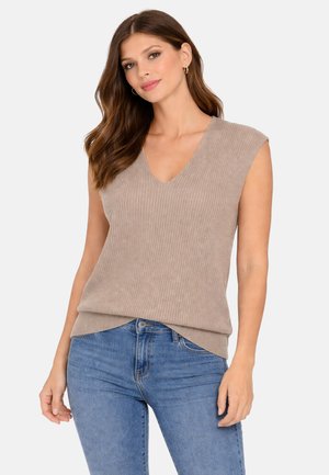 Donna che indossa un top smanicato beige a coste con scollo a V e jeans blu, in piedi davanti a uno sfondo bianco uniforme.