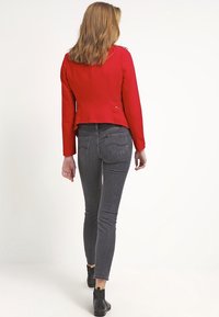 Veste ajustée rouge avec fermetures éclair, associée à un jean gris slim-fit. Des bottines noires à la cheville complètent la tenue. Tissu lisse, design structuré.