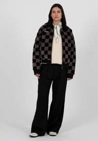 Frau trägt schwarze und braune karierte Jacke, cremefarbenen Pullover, schwarze weite Hosen, weißen Schal und weiße Schuhe und steht vor einem grauen Hintergrund.