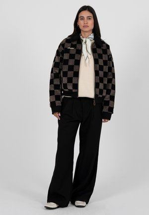 Žena v černohnědém kostkovaném saku, krémovém svetru, černých širokých kalhotách, bílém šátku a bílých botách stojící před šedým pozadím.