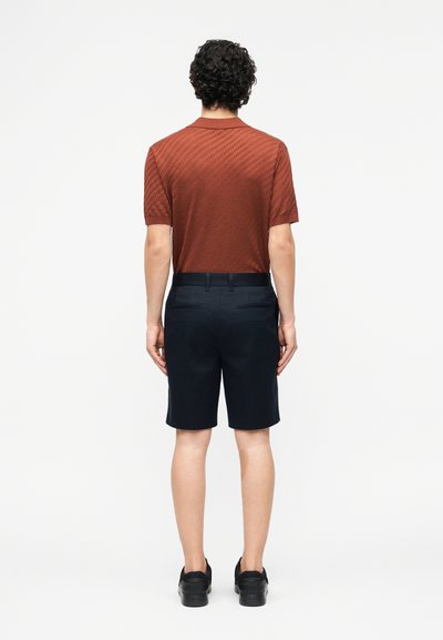 Άνδρας με σγουρά μαλλιά που φοράει πουκάμισο με κοντά μανίκια σε σκουριά χρώμα, σκούρα ναυτικά σορτς και μαύρα παπούτσια, στέκεται με την πλάτη προς τα εμάς σε λευκό φόντο.