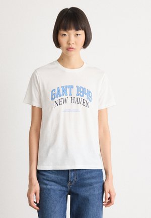 Kvinne med kort svart hår iført en hvit T-skjorte med teksten "GANT 1949 NEW HAVEN" og blå jeans, stående mot en hvit bakgrunn.