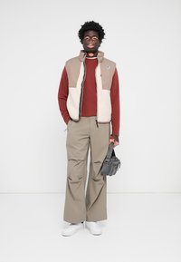 Beige and cream fleece vest over a maroon long-sleeve shirt, paired with wide beige trousers and white sneakers. Gray crossbody bag included.
