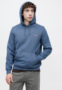 Μπλε φούτερ με κουκούλα από μαλακό ύφασμα, με μπροστινή τσέπη και μικρό λογότυπο στο στήθος. Φορεμένο πάνω από ένα σκούρο πουκάμισο.