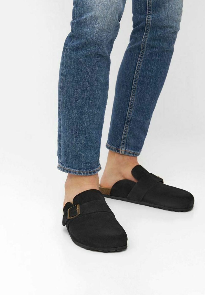 Svarta mocka träskor med rem och spänne, kombinerade med blå jeans, vilket visar en avslappnad design och texturerad yta.