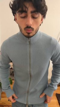 Νεαρός άνδρας με σκούρα μαλλιά και γένια φοράει ανοιχτό γαλάζιο πλεκτό πουλόβερ με φερμουάρ, στέκεται μέσα σε κλειστό χώρο με τα χέρια κοντά στις τσέπες.